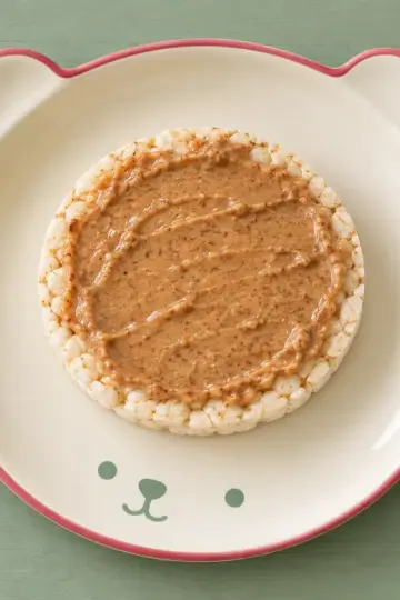 Reiswaffel mit Mandelmus auf cremefarbenem Keramikgeschirr mit himbeerfarbenem Rand auf salbeifarbenem Holz.