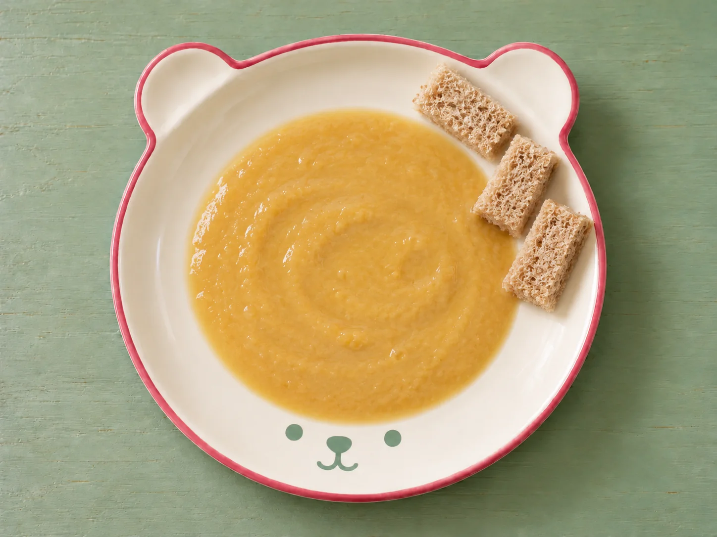 Goldgelbe Linsensuppe ohne Gemüse mit weichen Vollkornbrotstreifen auf einem cremefarbenen Bärenteller mit himbeerfarbenem Rand.